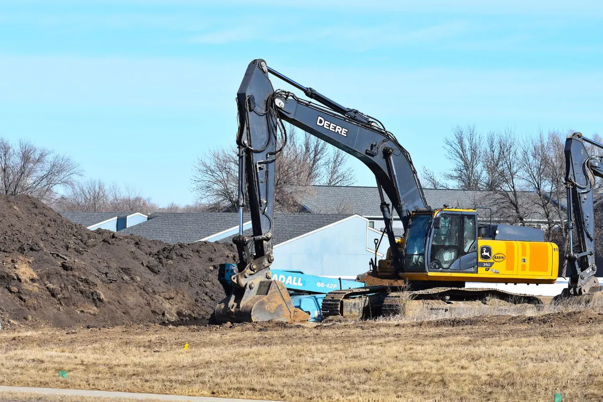 Excavator working beside dirt piles on a residential lot with houses behind